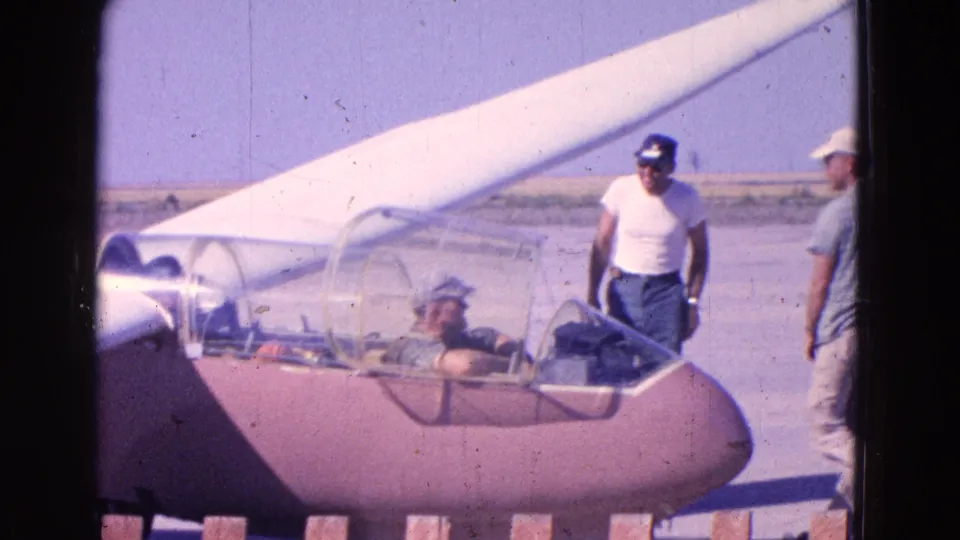 Two men stand near the side of the glider while a pilot sits under the clear canopy, suggesting informal preflight activity.
