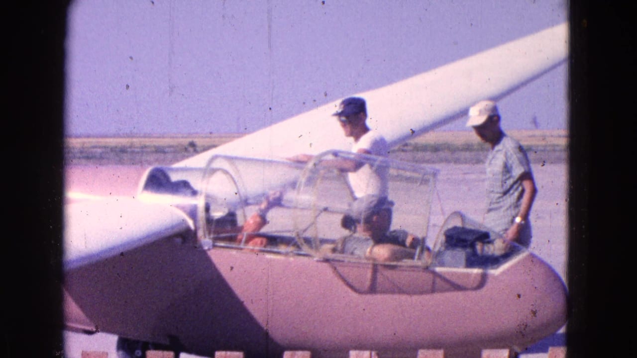 Men stand beside a pink glider with an open clear canopy on a runway.