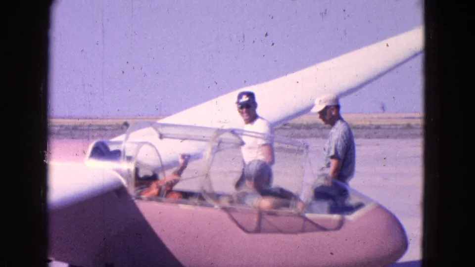 Home-movie frame from the 1964 Washington DC-area clip showing a glider cockpit, open canopy, seated pilot, and two men nearby.