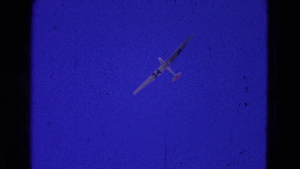 1964 8mm frame showing a small glider marked with the number 34 against a saturated blue-purple sky, with visible grain and scratches.