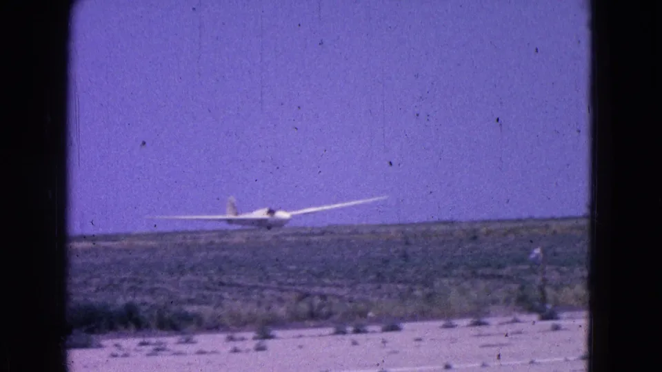 1964 8mm frame showing a glider low over a flat arid field, with sparse vegetation, pale foreground ground, and a purple-blue sky.