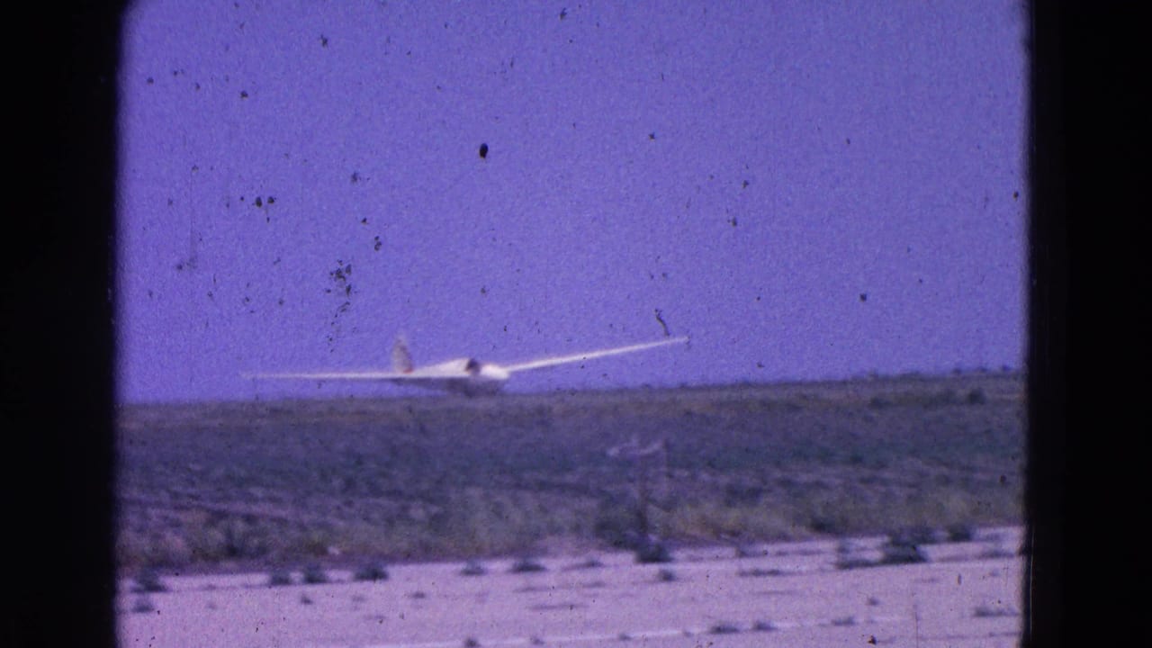 White glider low over a sparse airfield beneath a purple-blue sky.
