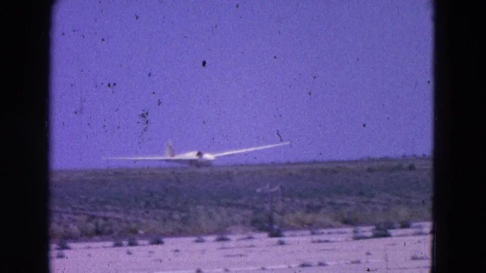 1964 8mm frame of a white glider nearing a low, scrubby airfield horizon, with visible film grain and dark frame edges.