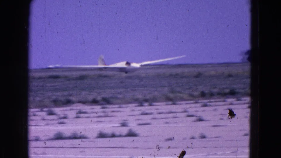 1964 8mm view of a white glider moving low across an open airfield, with sandy foreground and visible film wear.
