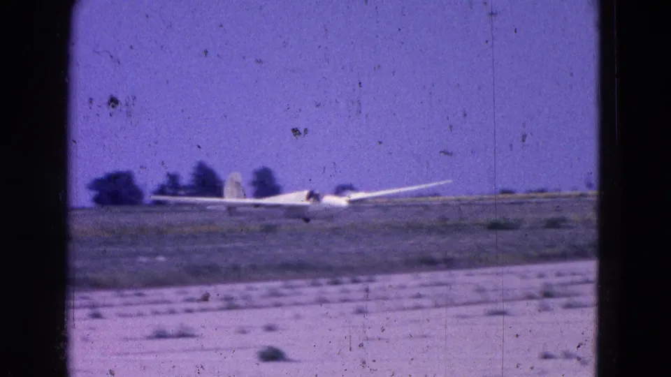 White glider appears at the end of its landing run near a flat airfield, with distant dark trees and 8mm dust visible.