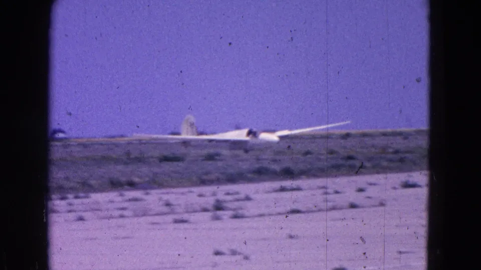 1964 8mm frame of a white glider crossing a dusty airfield, with dark film borders and visible surface specks.