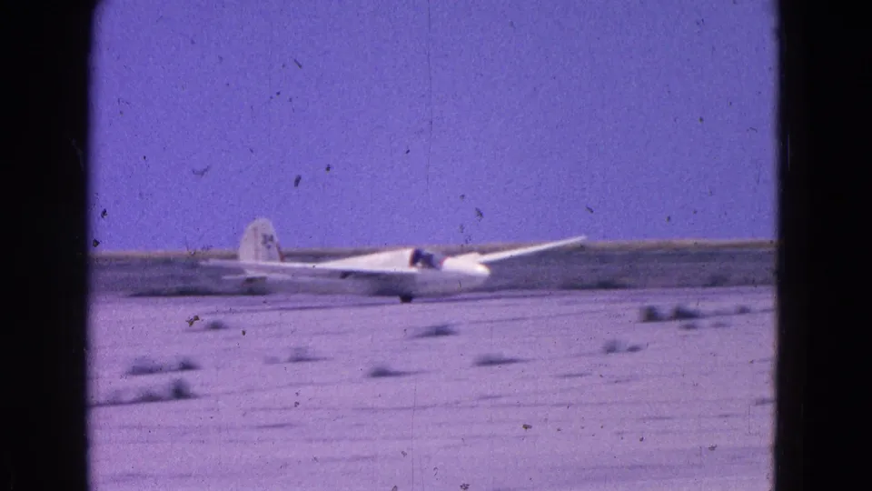 1964 home-movie still showing the white glider low on the dusty airfield, with grain and dark 8mm frame edges.