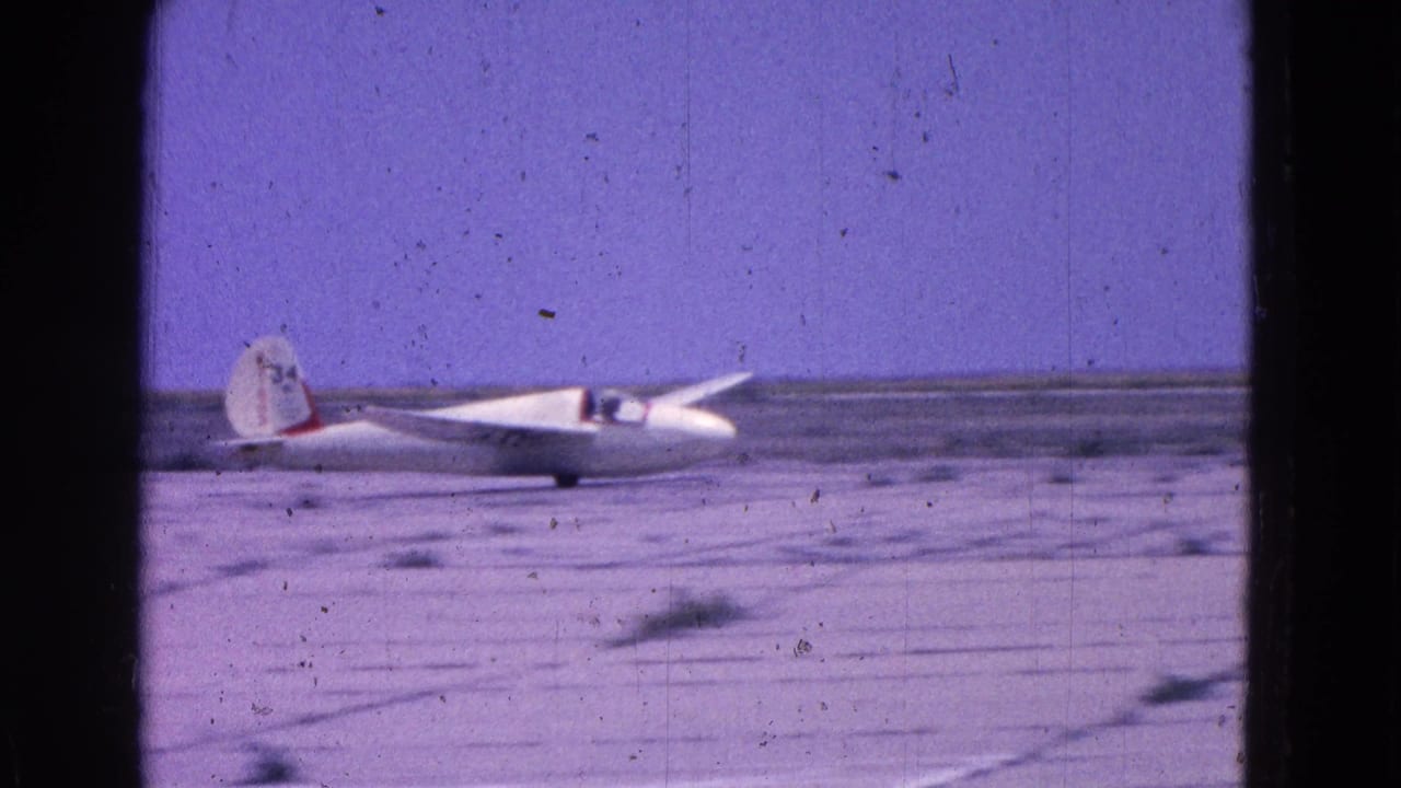 Small white glider on a flat airfield with a pilot visible in the cockpit.