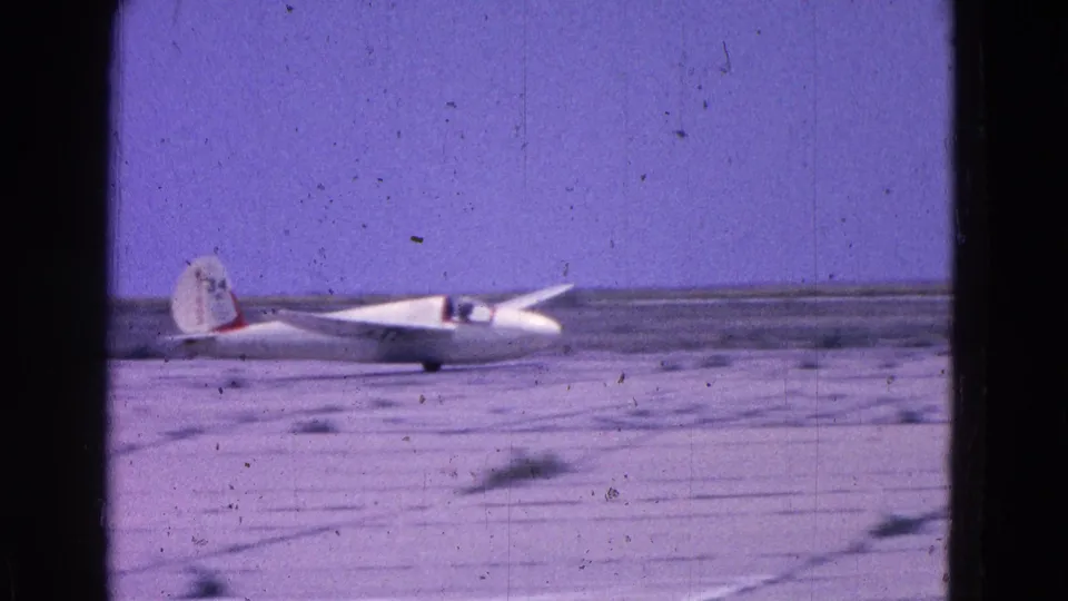 1964 8mm frame of a white experimental glider on a flat Washington DC airfield, with red tail markings and a pilot visible.