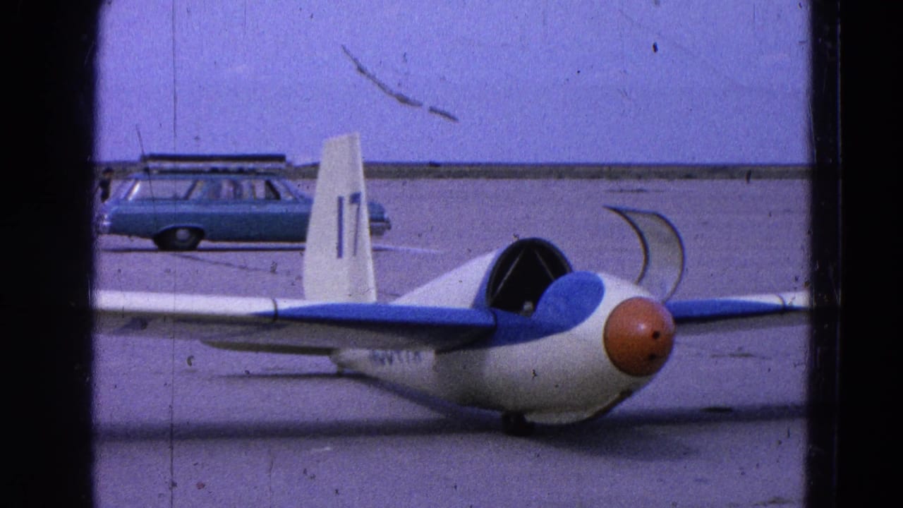 Small white-and-blue glider with orange nose on an airfield, with a station wagon behind it.