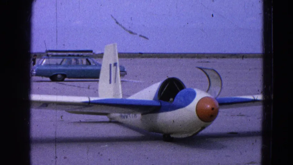 A small white-and-blue glider with an orange nose sits on open pavement at a 1964 Washington, DC area airfield, with a period station wagon behind it.