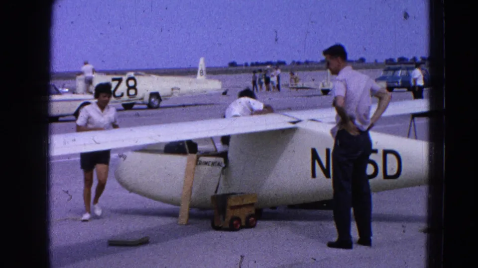 People in casual 1960s clothing gather around the wing and fuselage of a white glider marked N65D at a Washington, DC area airfield.