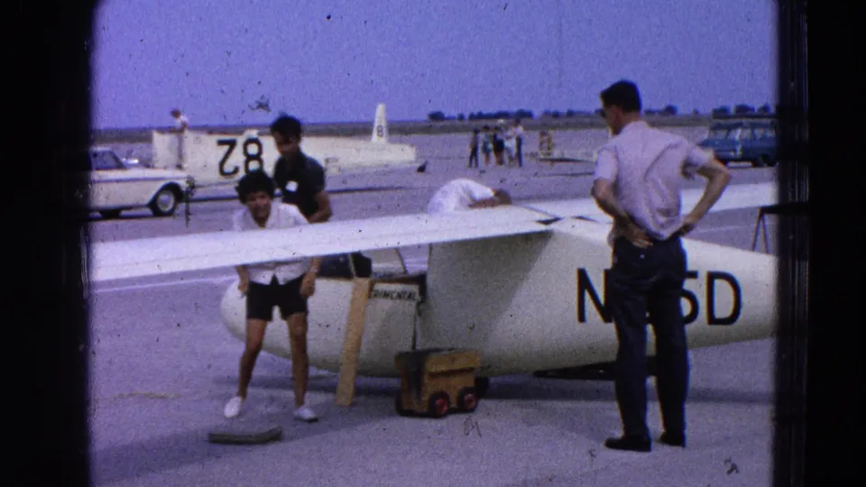 Several adults stand beside a white glider marked N65D on a 1964 Washington, DC area airfield, with other sailplanes and parked cars in the distance.