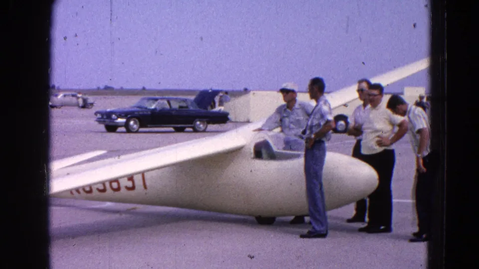 1964 Washington, DC home-movie frame showing men inspecting a white glider on an airfield, with a dark 1960s sedan in the background.
