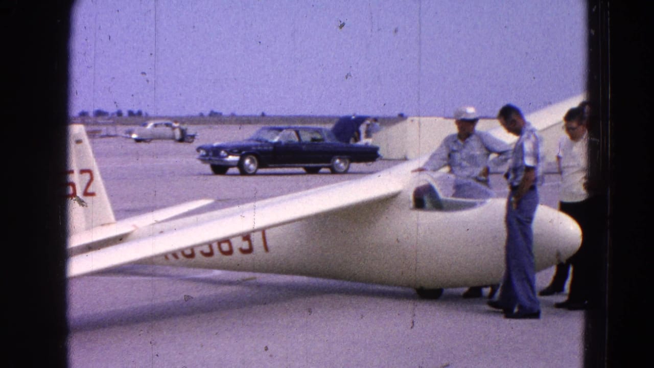 Men stand near the glider cockpit and nose, with parked cars across the airfield.