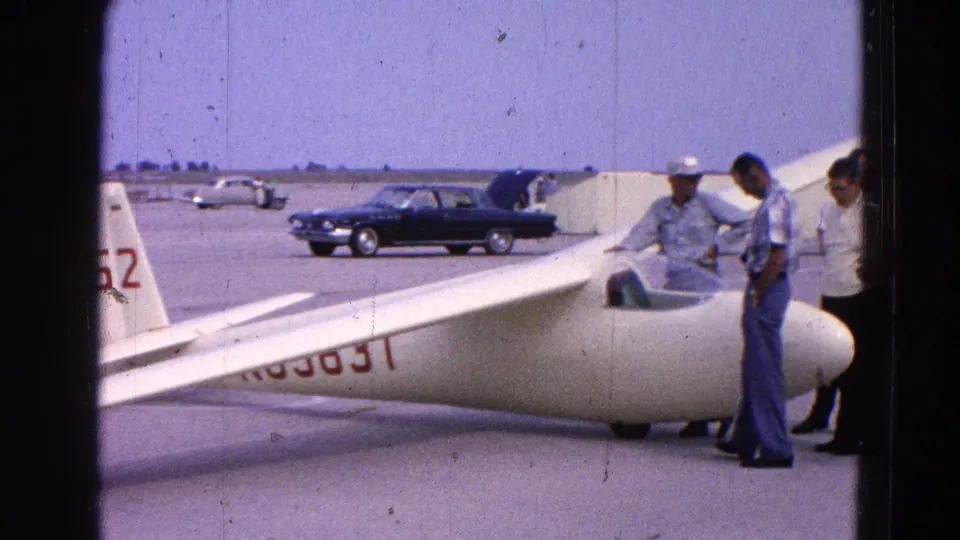 Men in casual 1960s clothing appear to inspect a white glider at a Washington, DC airfield, with parked cars and open pavement behind them.