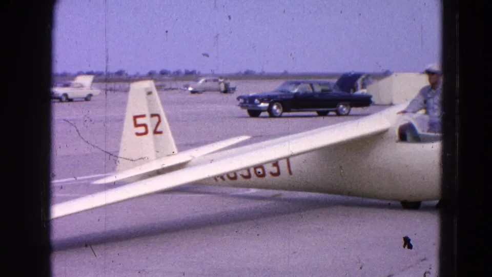 The glider tail marked 52 and part of the fuselage registration are visible on the airfield, with parked cars and a dark sedan beyond.