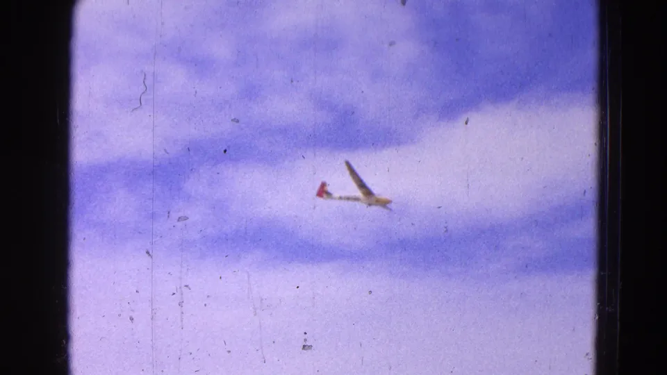A distant glider is seen in flight against a cloudy sky, continuing the Washington, DC 1964 airfield sequence from ground inspection to flight.