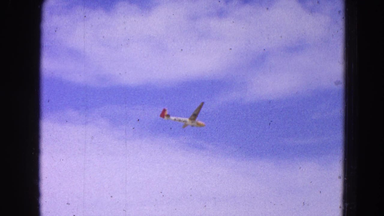 Distant glider flying in a cloudy sky with visible film scratches and dust.