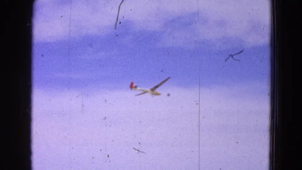 A small glider descends through a pale cloudy sky, seen in soft-focus 8mm home-movie footage.