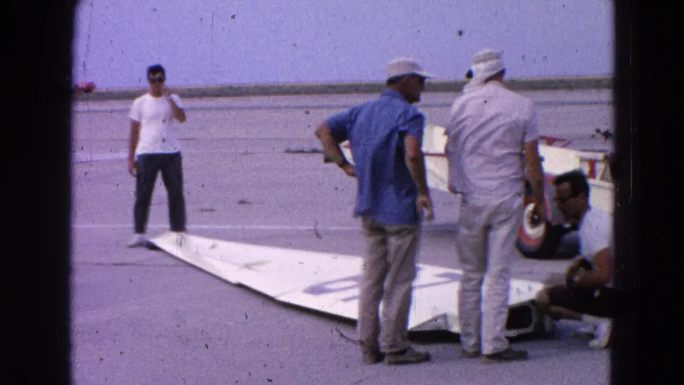 Several men in casual clothing stand and crouch beside a detached glider wing on a 1964 Washington-area airfield.