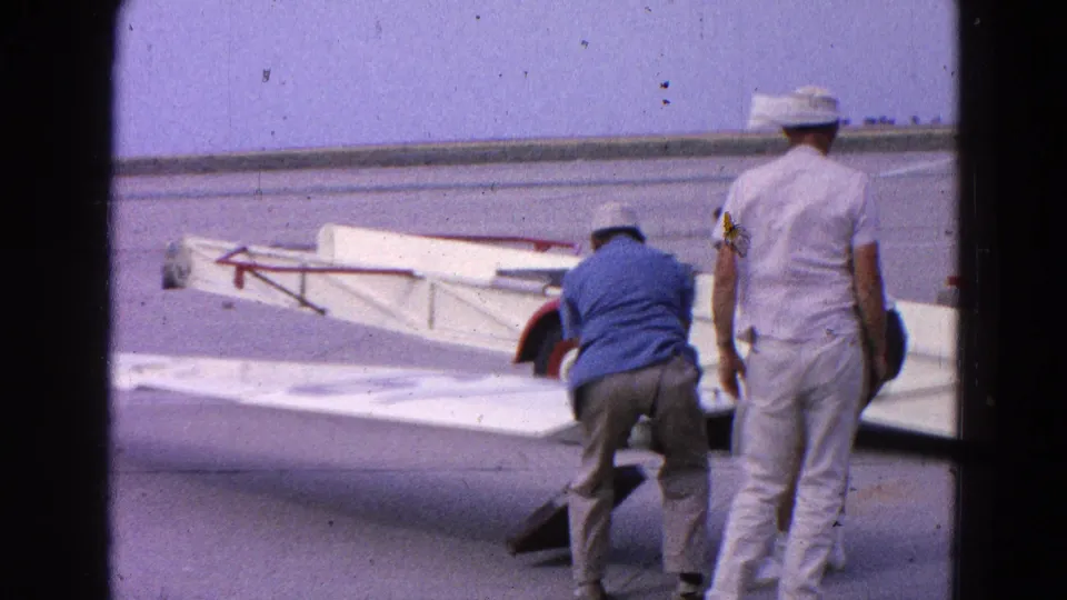 Two men stand near the white glider and wing on the tarmac, with 8mm film grain and gate edges visible in the frame.