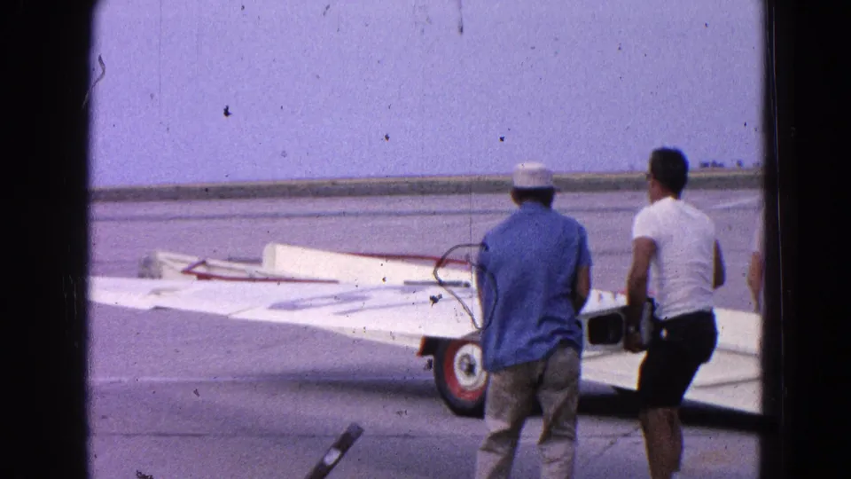 In 1964 Washington DC footage, two men guide a white airplane wing on a trailer across open tarmac.