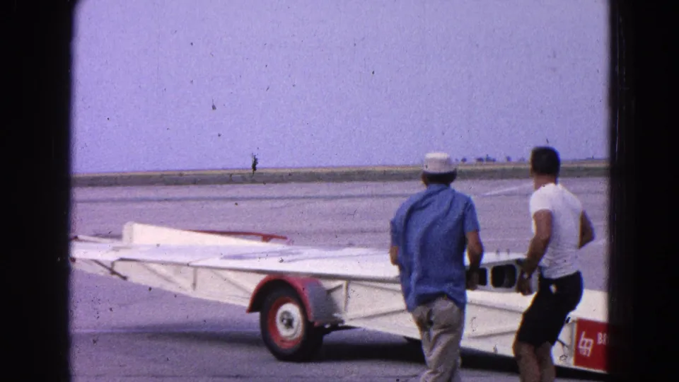 Two men move alongside a large white airplane wing and red-wheeled trailer on an open Washington DC airfield.