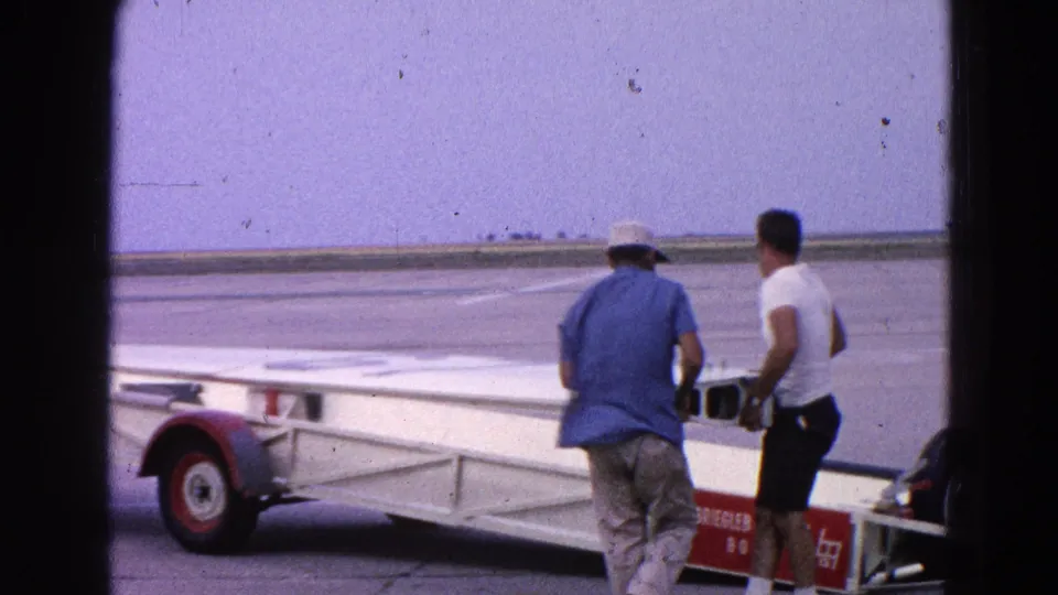 The men continue maneuvering the white airplane wing on its trailer across the tarmac in 1964 Washington DC footage.