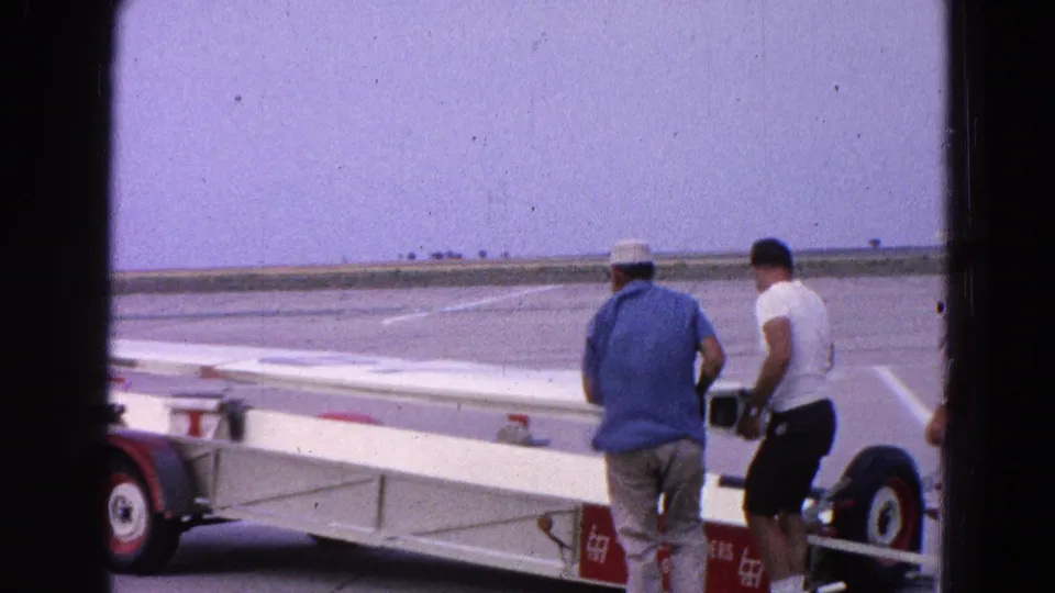 Two men guide a long white airplane wing on a small trailer across marked airfield pavement under a pale sky.