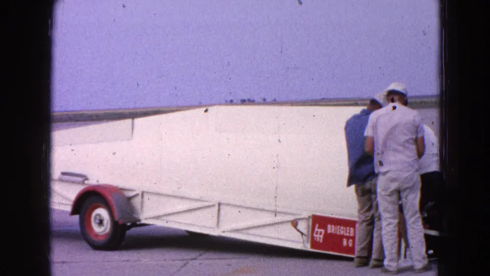 1964 home-movie frame near Washington, DC, showing men inspecting a white glider on a trailer at a flat airfield.