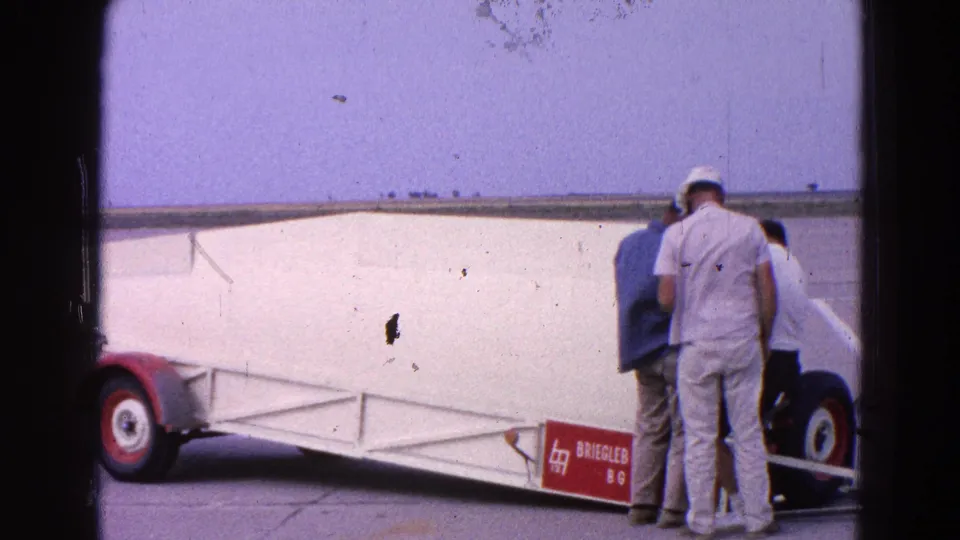 1964 airfield footage shows a white glider on a trailer with several people standing close to the fuselage.