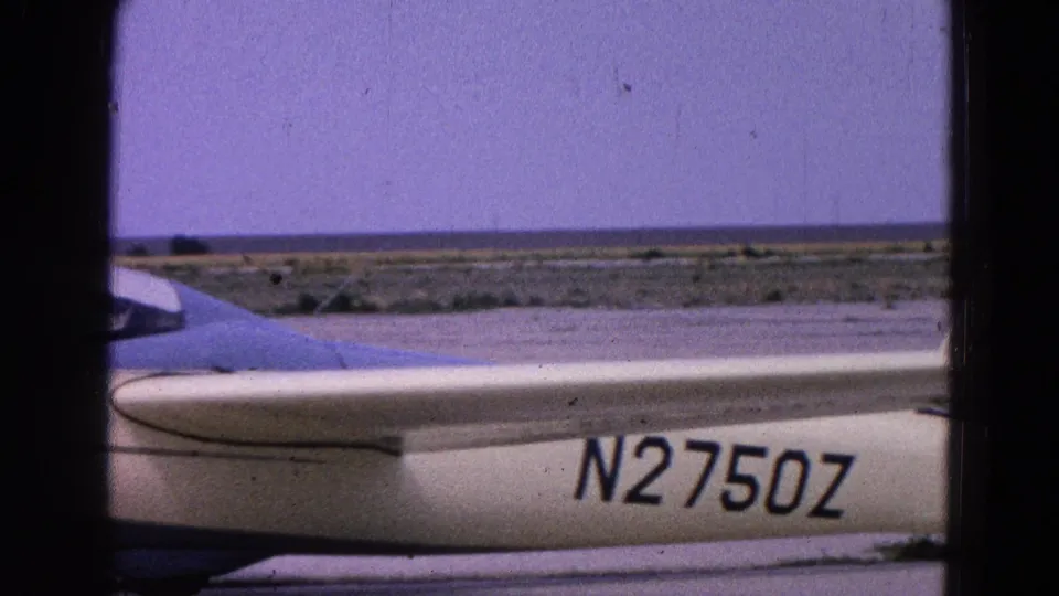 1964 8mm frame of a white glider on an open airfield, with registration N2750Z visible and flat landscape beyond.