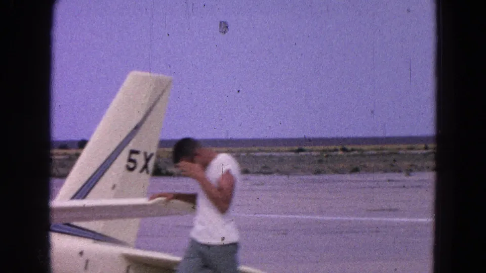 1964 8mm frame showing a man beside the white glider tail marked 5X, with tarmac and open landscape behind him.