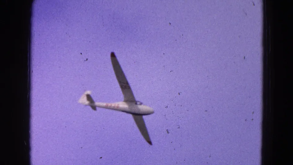 1964 8mm frame of the glider airborne against a purple-blue sky, seen from below at a distance.