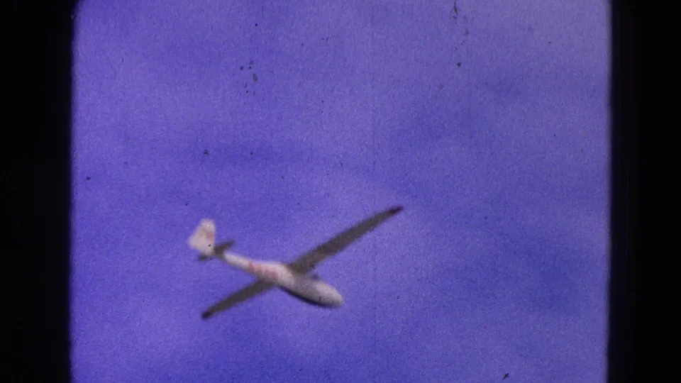 A white glider with dark wings passes through the lower frame, set against layered blue clouds in 1964 home-movie footage.