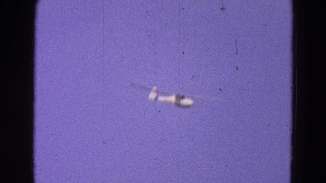 Small white glider flying through a purple-blue sky with film grain and dark frame edges.