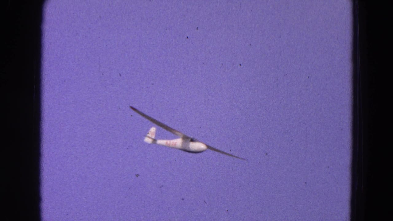 White glider banking in open sky with red markings visible on the tail area.