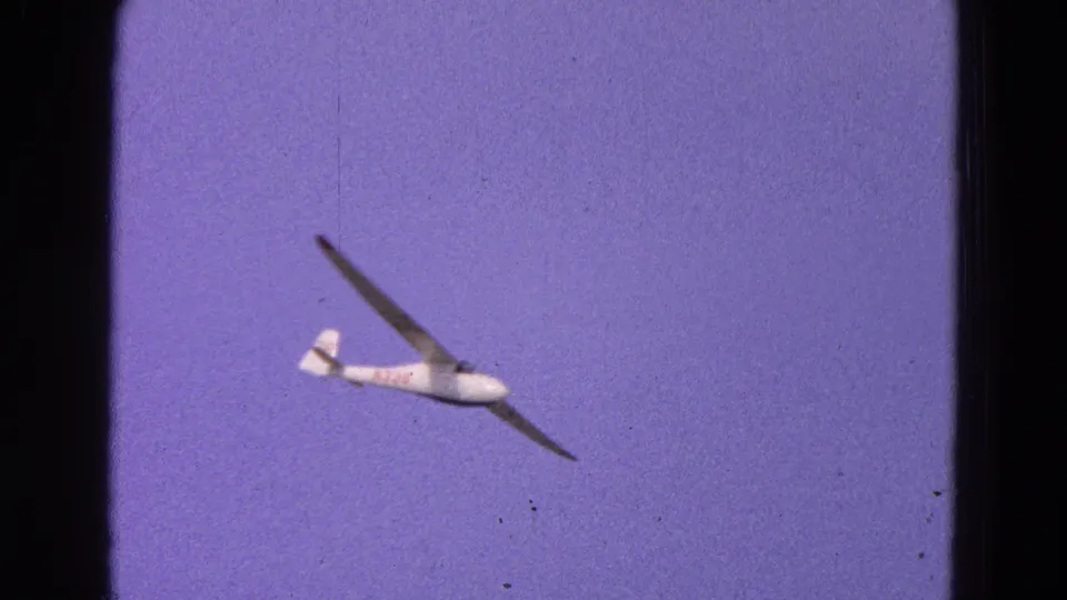 1964 home-movie view of a white glider banking overhead, its dark wings silhouetted against a purple-blue sky.