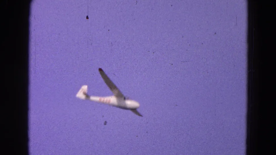 White glider with red markings passes through the frame in 1964 Washington, DC 8mm footage, softened by focus and film grain.