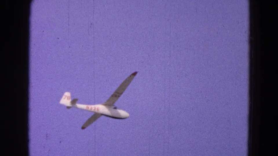 1964 8mm frame showing the white glider banking, with red markings and tail number 78 visible in a purple-blue sky.