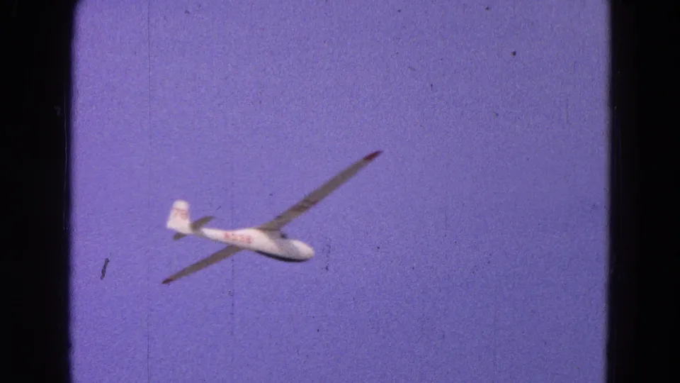 1964 8mm frame of the white glider banking away, with visible film grain, dust, and saturated purple-blue sky.