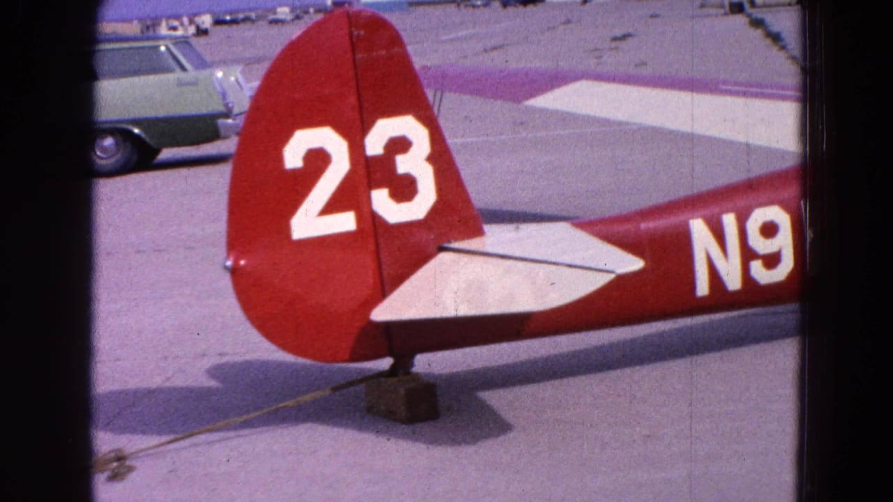 Red aircraft tail with white number 23 beside airport pavement and a parked station wagon.