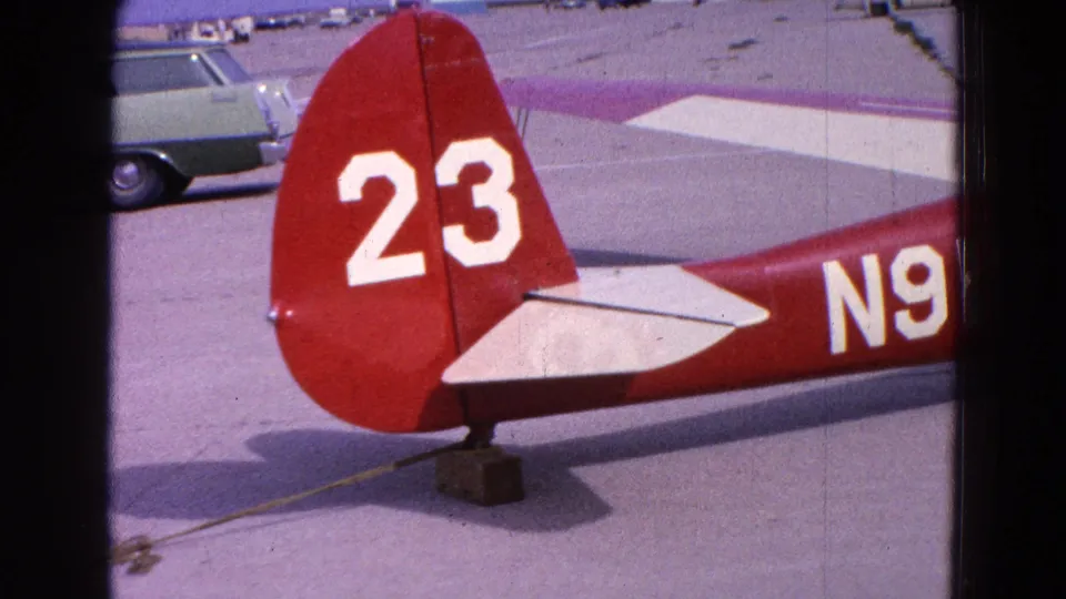1964 Washington, DC home-movie frame showing the tail of a red-and-white aircraft marked 23, with airport pavement and a parked station wagon behind it.