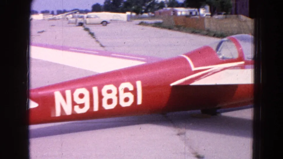 1964 Washington, DC home-movie frame showing the aircraft side with registration N91861, white wing, tarmac, parked vehicles, and distant low buildings.