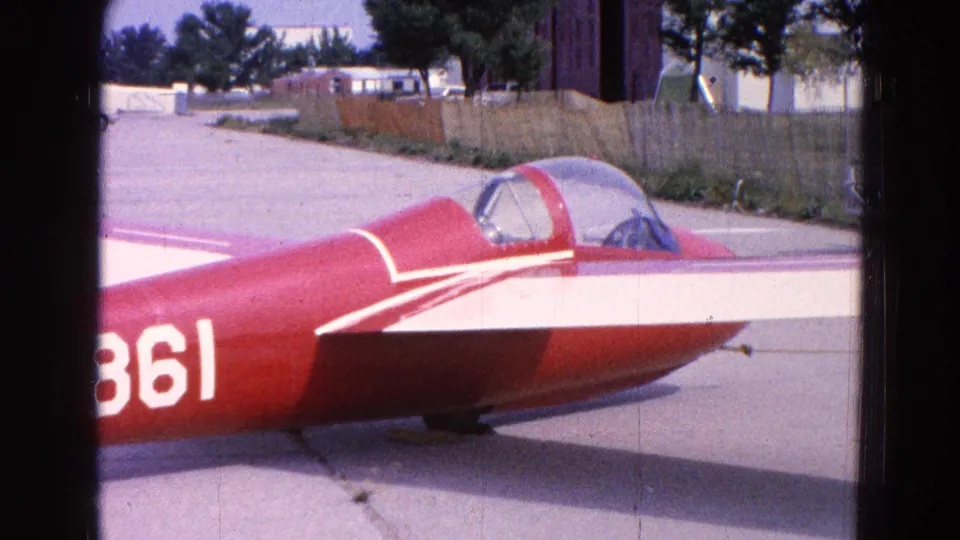 1964 Washington, DC frame showing the red-and-white aircraft cockpit canopy, side stripe, tarmac, fence line, trees, and low buildings in the background.