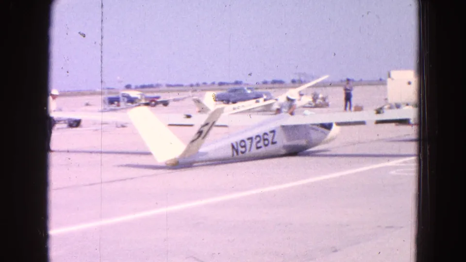 The white glider remains parked on the airfield tarmac, with N9726Z visible, distant small planes, cars, and a person near the background.