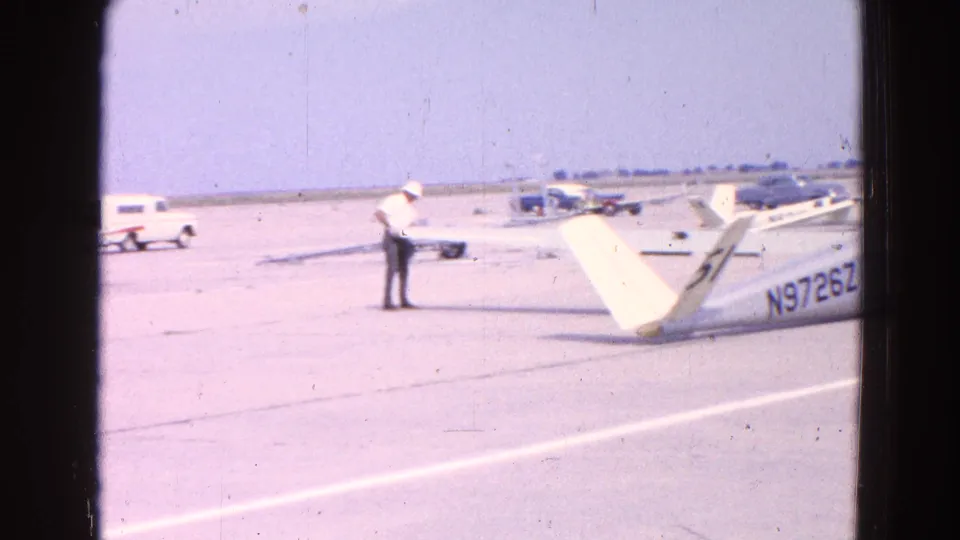A man in a white hat stands on the tarmac near the glider tail, with small aircraft and vehicles spread across the airfield behind him.