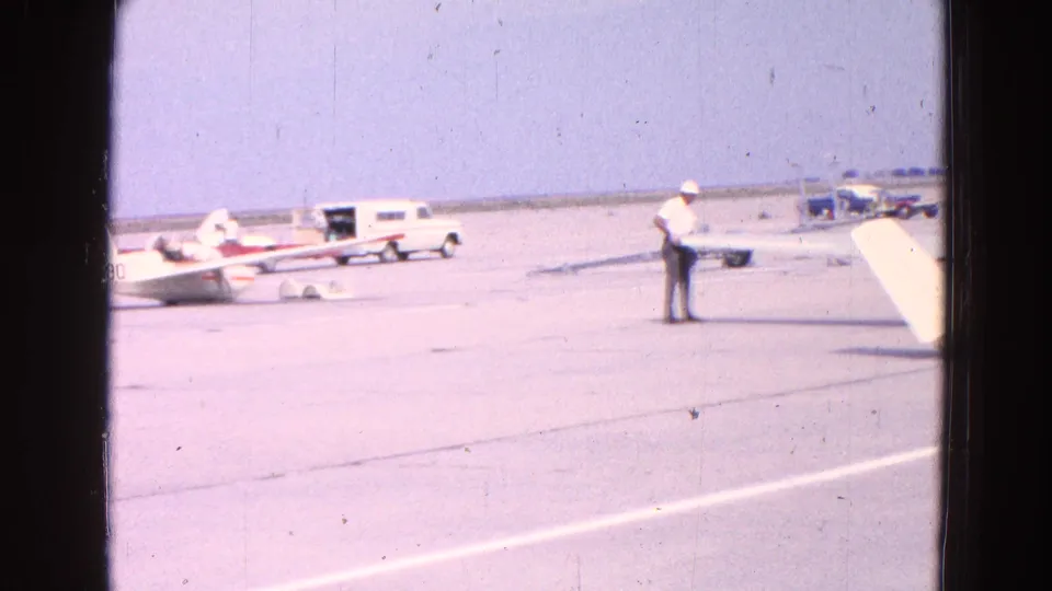 A man in a white hat stands on the airfield tarmac near a red and white glider, with another small plane and vehicles visible in the background.