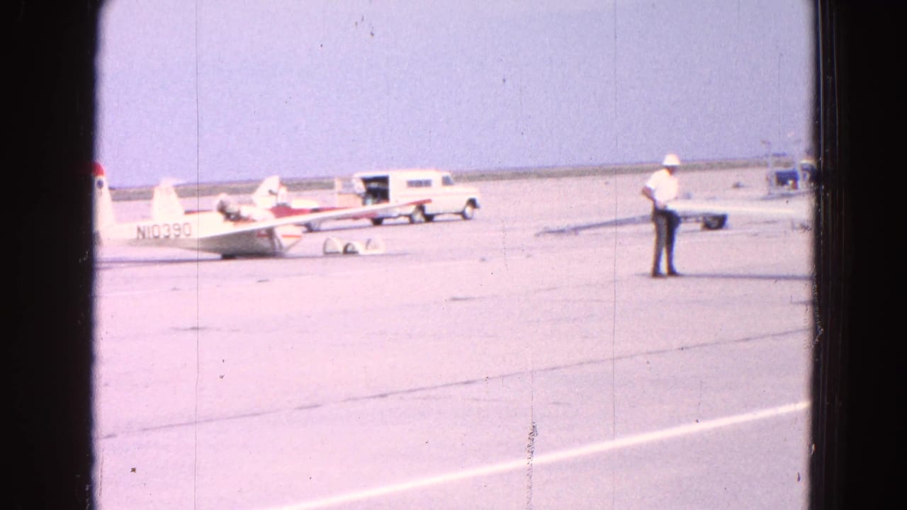 Small white aircraft on a sunlit tarmac with a man standing nearby and a white van in the background.